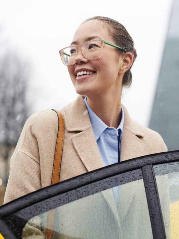 Portrait of young pretty  Asian businesswoman getting in  taxi cab on rainy autumn street, smiling and looking away while opening car door